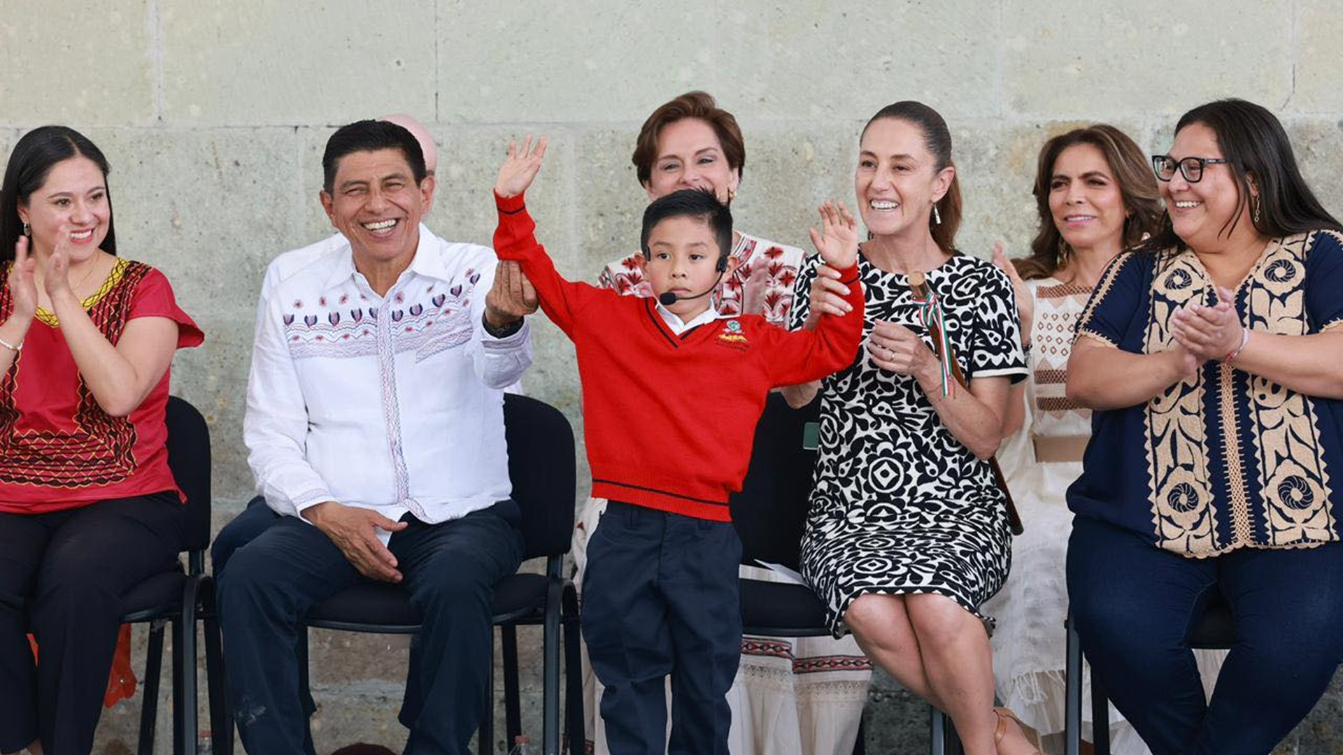 Cinco mujeres y un hombre junto a un niño con suéter rojo que levanta las manos.