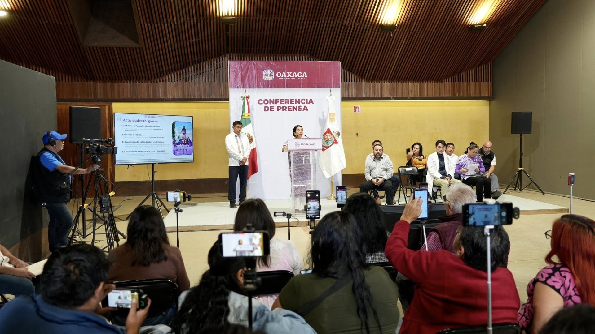 Periodistas observan el desarrollo de una conferencia de prensa en la que habla una mujer con traje tipico indígena.