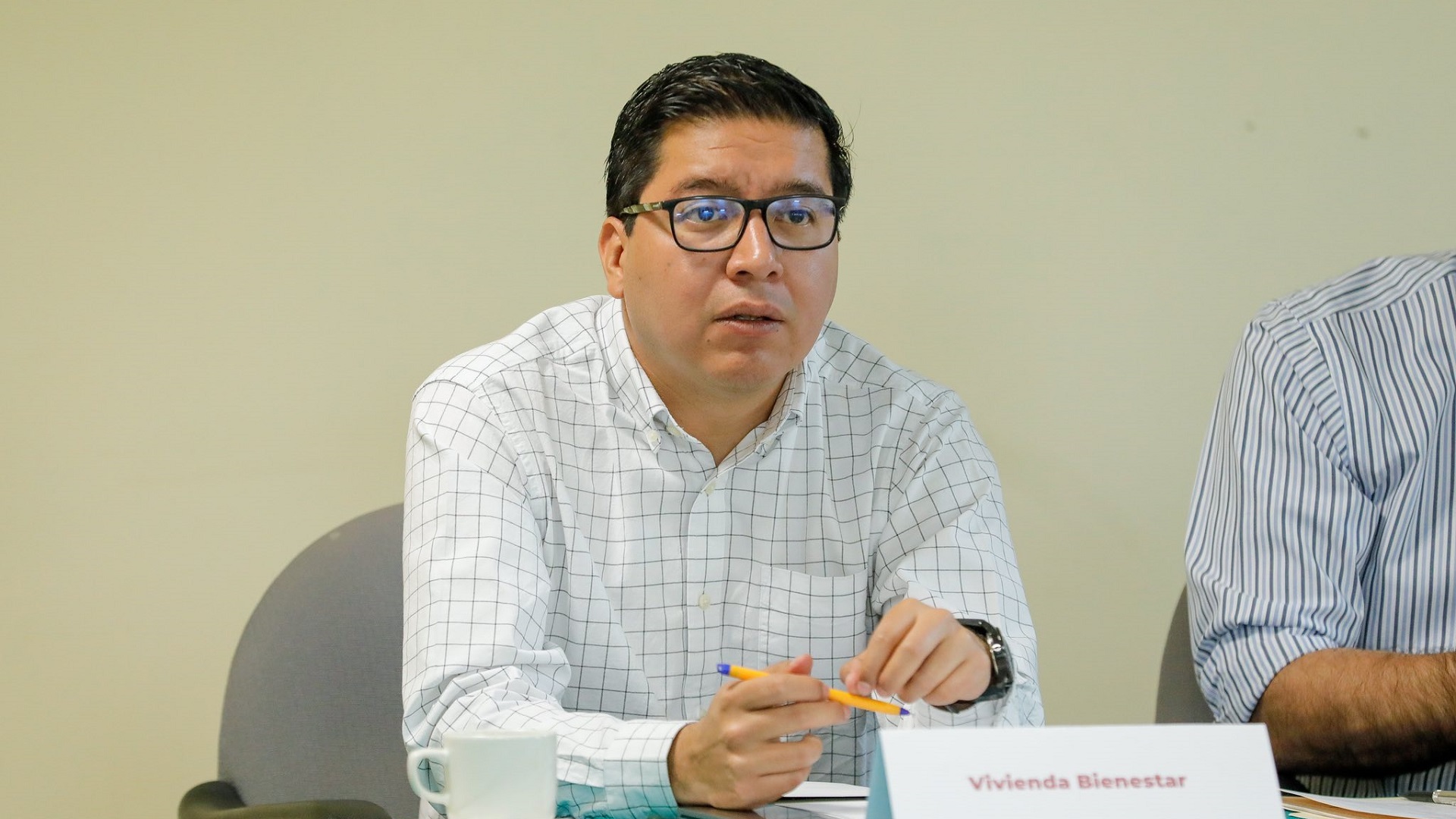 Hombre con camisa blanca y lentes en un escritorio y un papel con la leyenda "Vivienda Bienestar".