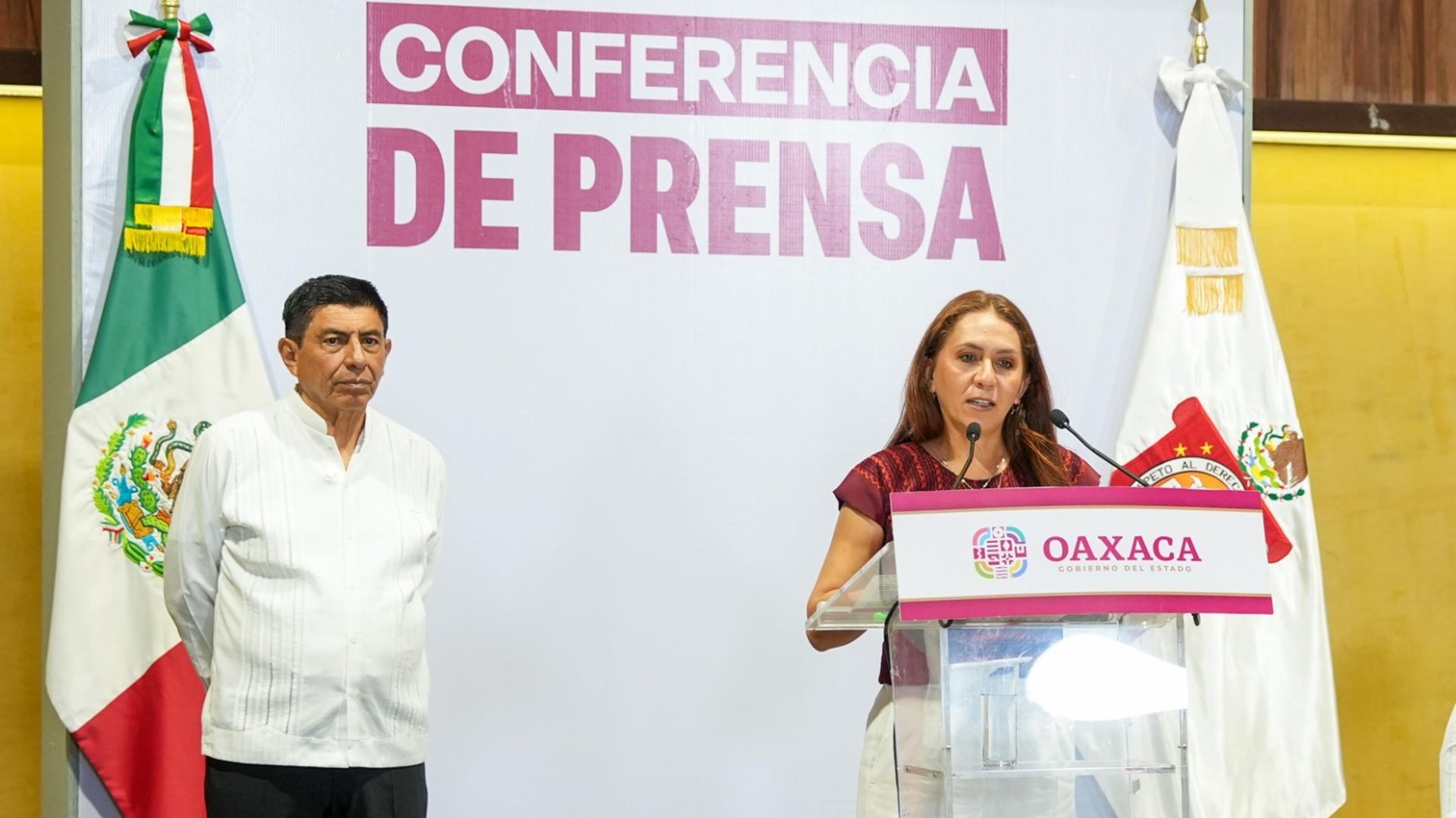 Sildia Mecott parada en un estrado da una conferencia de prensa, con el Salomón Jara de pie atrás de ella en la conferencia de prensa.