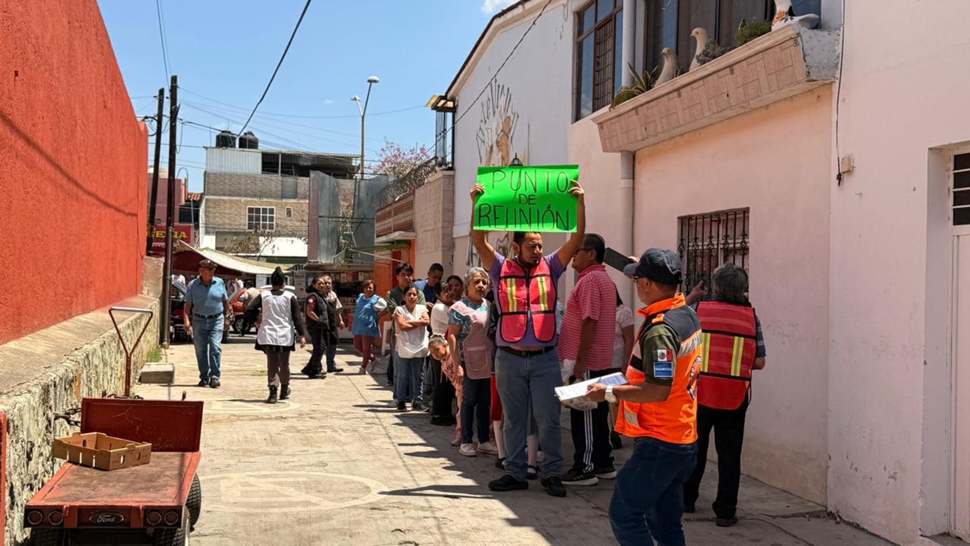 Un hombre con chalero naranja sostiene un carten con la leyenda "Punto re reunión", en un simulacro de sismo; está rodeado por varias personas más.