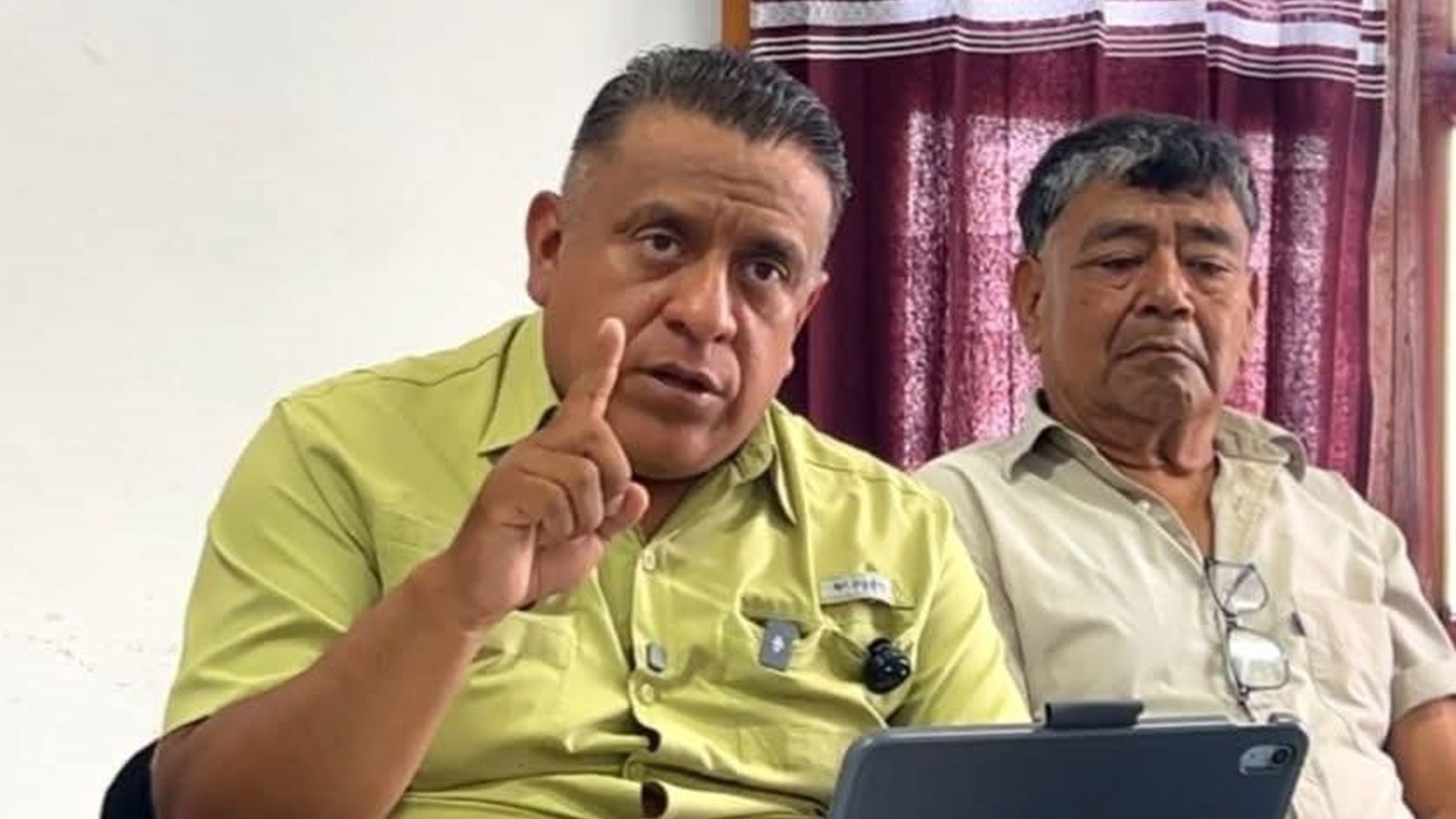 Esaú López con camisa verde sentado en una mesa y levantando la mano, junto a un hombre callado y serio.