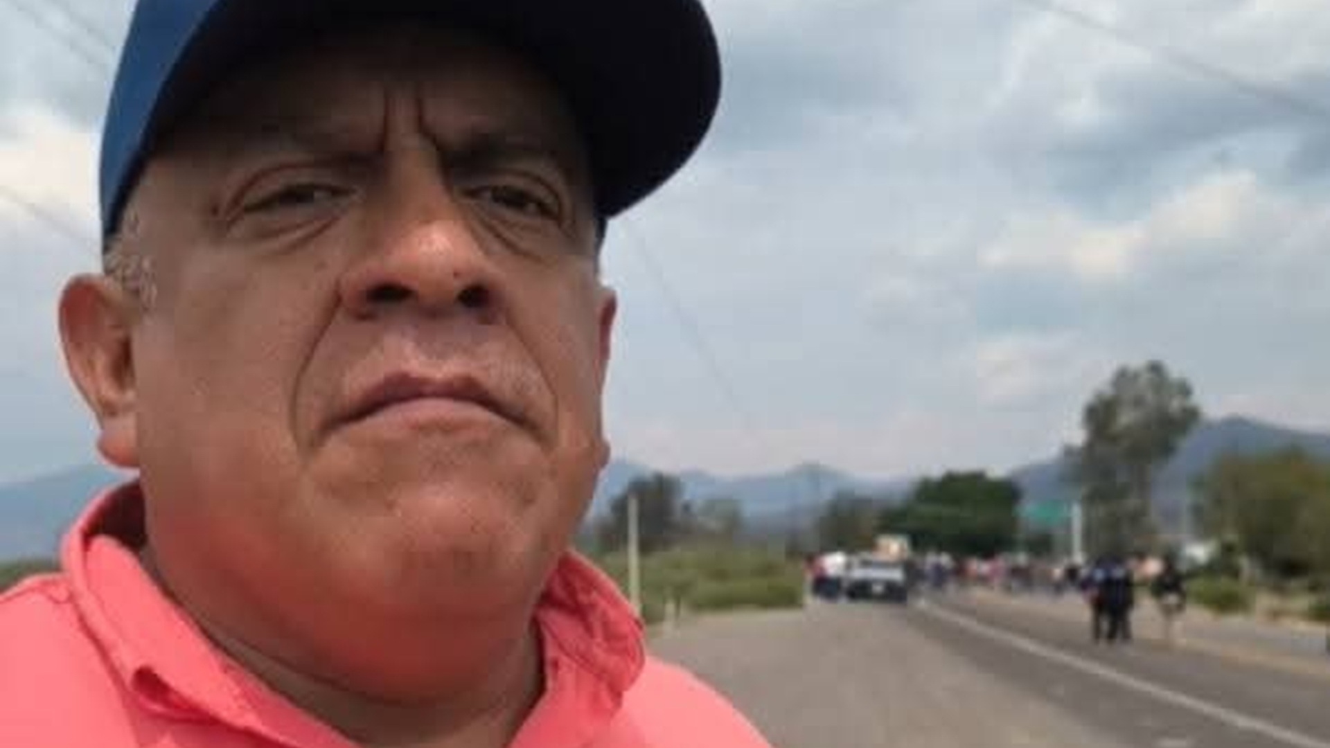 Esaú López con playera narajna y gorra en una carretera con vehículos al fondo.
