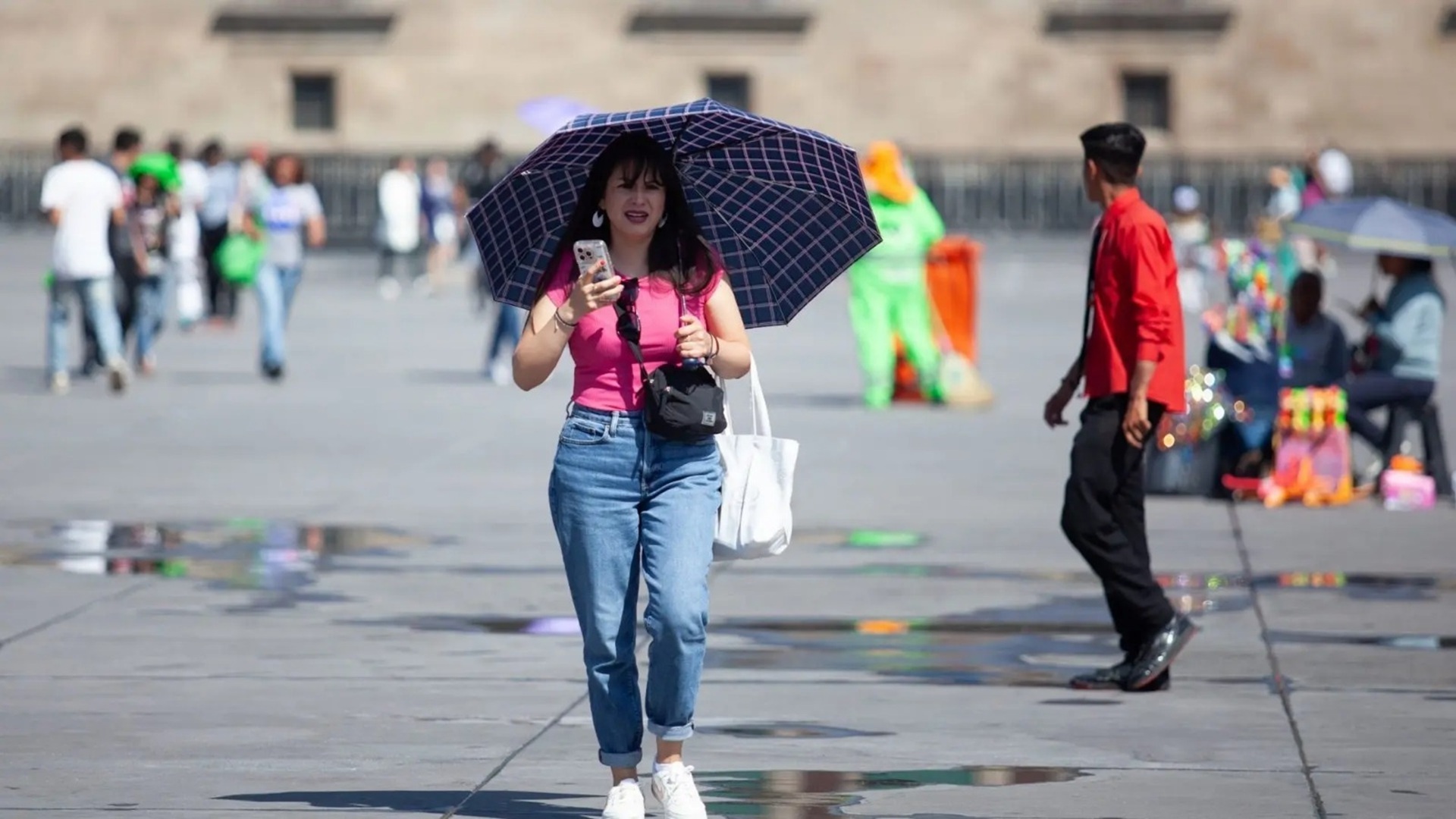 Una mujer con una sombrilla y cargando bolsas, junto a más personas en una plaza pública.