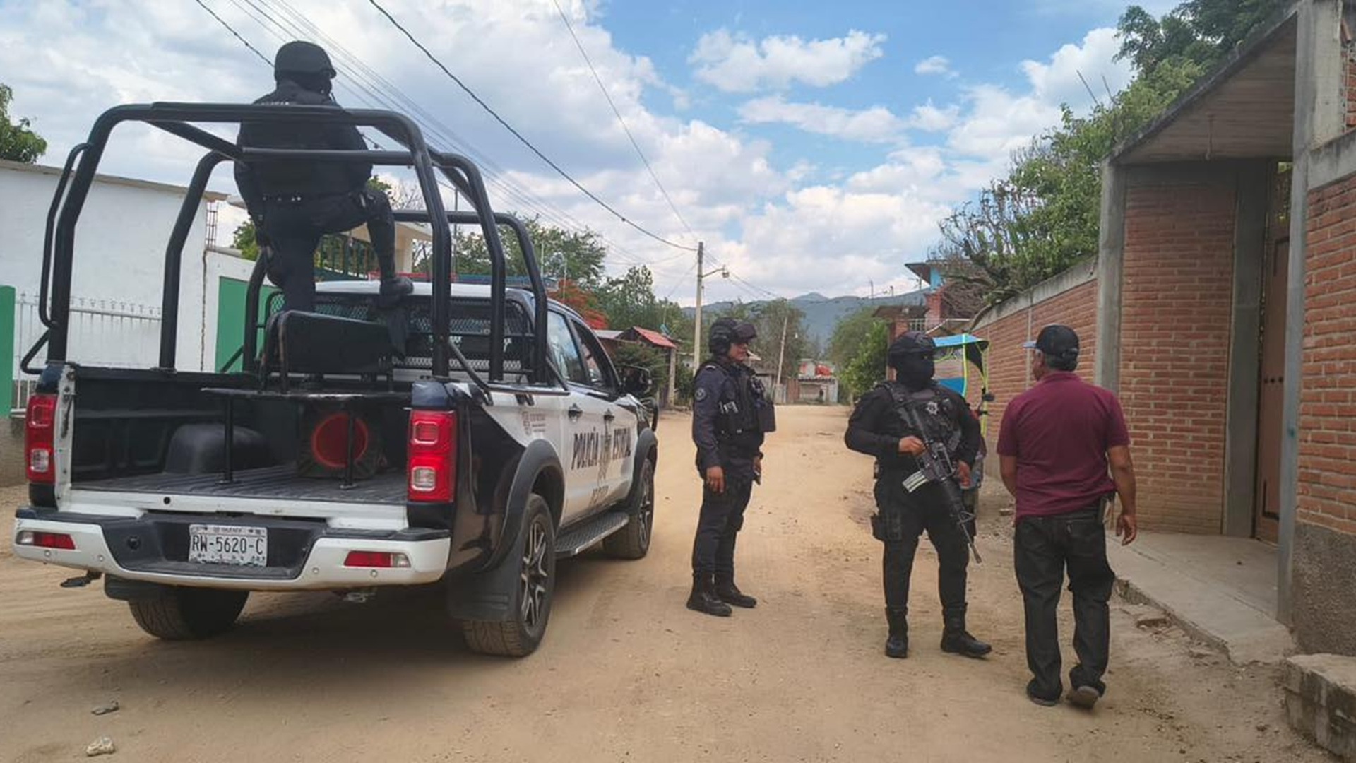 Policías armados junto a una patrulla, aparentemente interrogando a un hombre de camisa roja en una calle de terracería.
