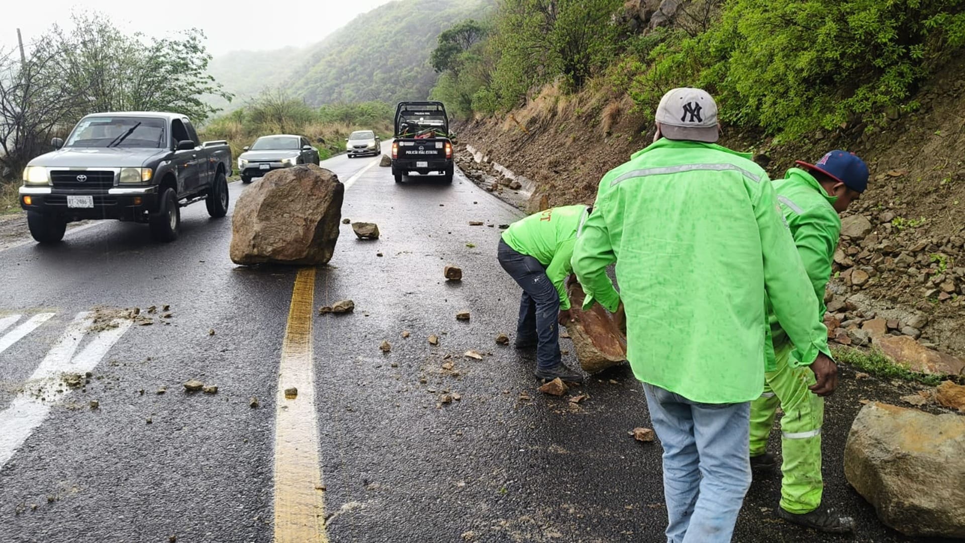 Varias personas con camisolas verdes remueven grandes rocas sobre una carretera, donde además hay varios autos detenidos.