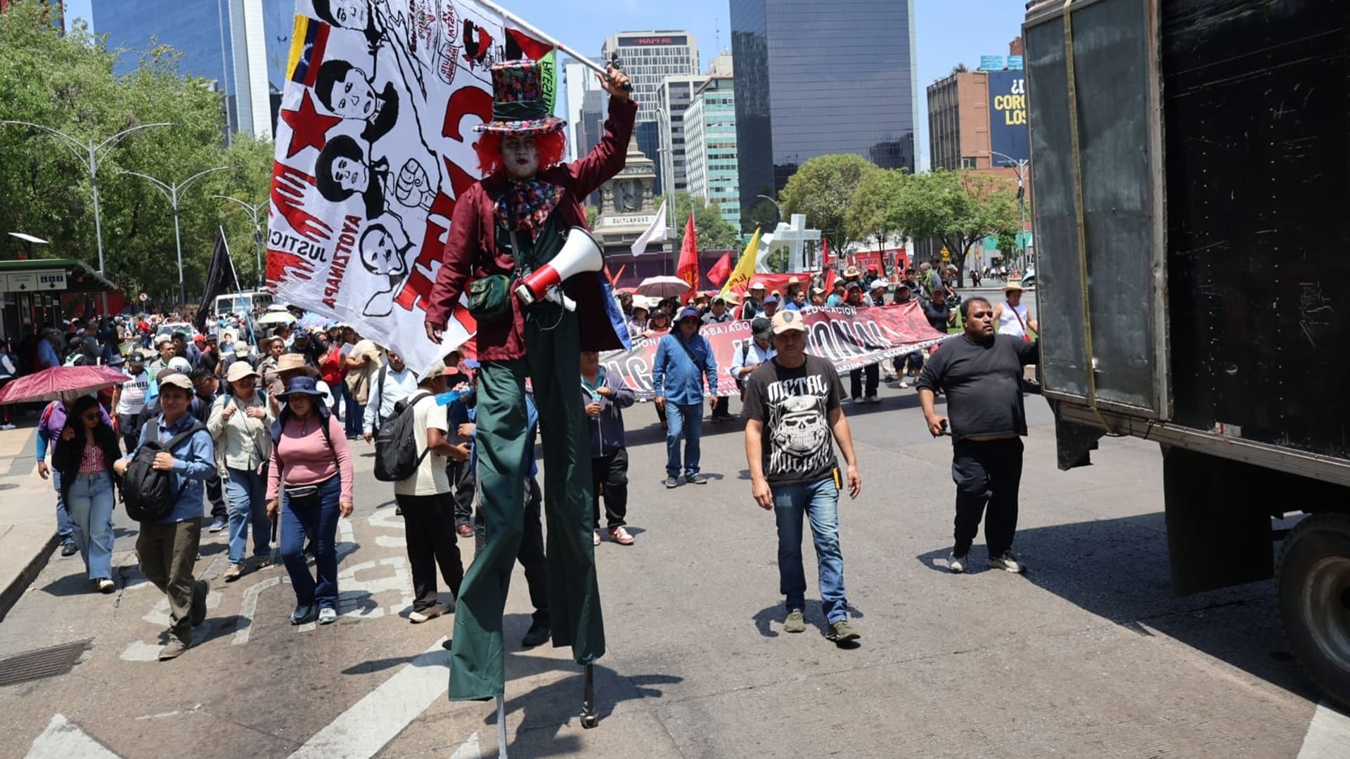 Decenas de personas marchan en una calle entre sombrillas y banderas, junto a una camioneta.