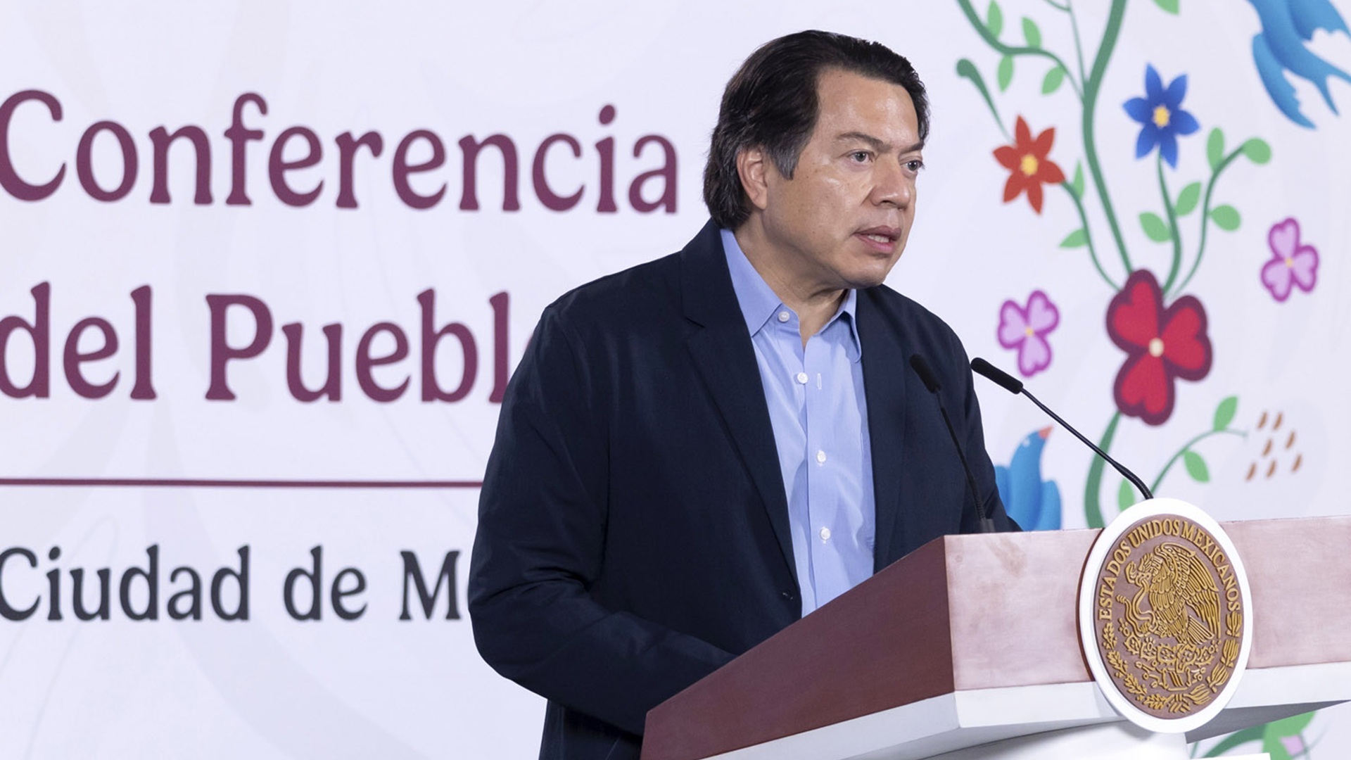 Mario Delgado con traje oscuro y camisa azul habla desde un atril con micrófono.