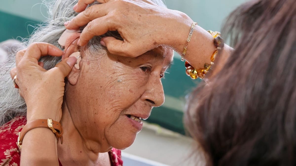 Una mujer coloca a otra mujer adulta mayor un aparato auditivo en la oreja derecha
