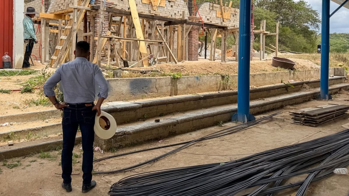 Un hombre con jeans y sombrero en mano supervisa junto a unas varillas una obra de construcción donde se ve un trabajador 