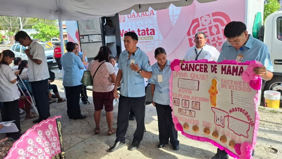 Hombres de Servicios de Salud de Oaxaca muestran cartulinas alusivas al cáncer de mama a mujeres que reciben capacitación en una calle frente a una unidad móvil