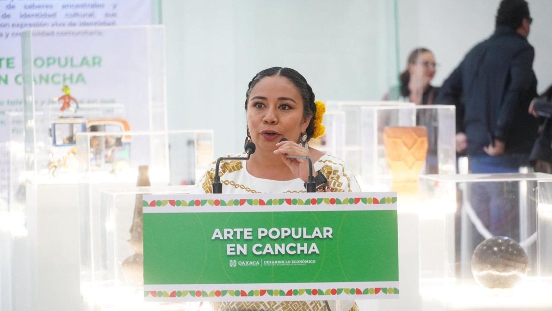Una mujer da un discurso con un micrófono y rodeada de piezas en exhibición.