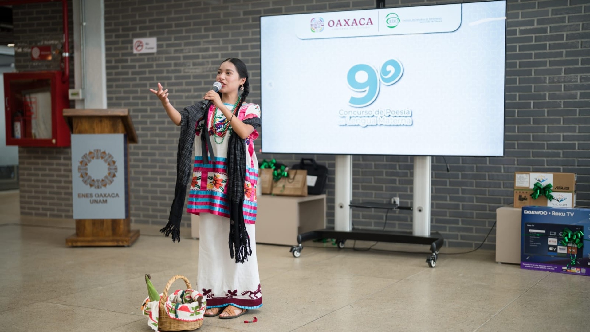 Una mujer joven con ropa regional oaxaqueña y un micrófono en la mano junto a una canasta y hablando a un auditorio que no se ve.
