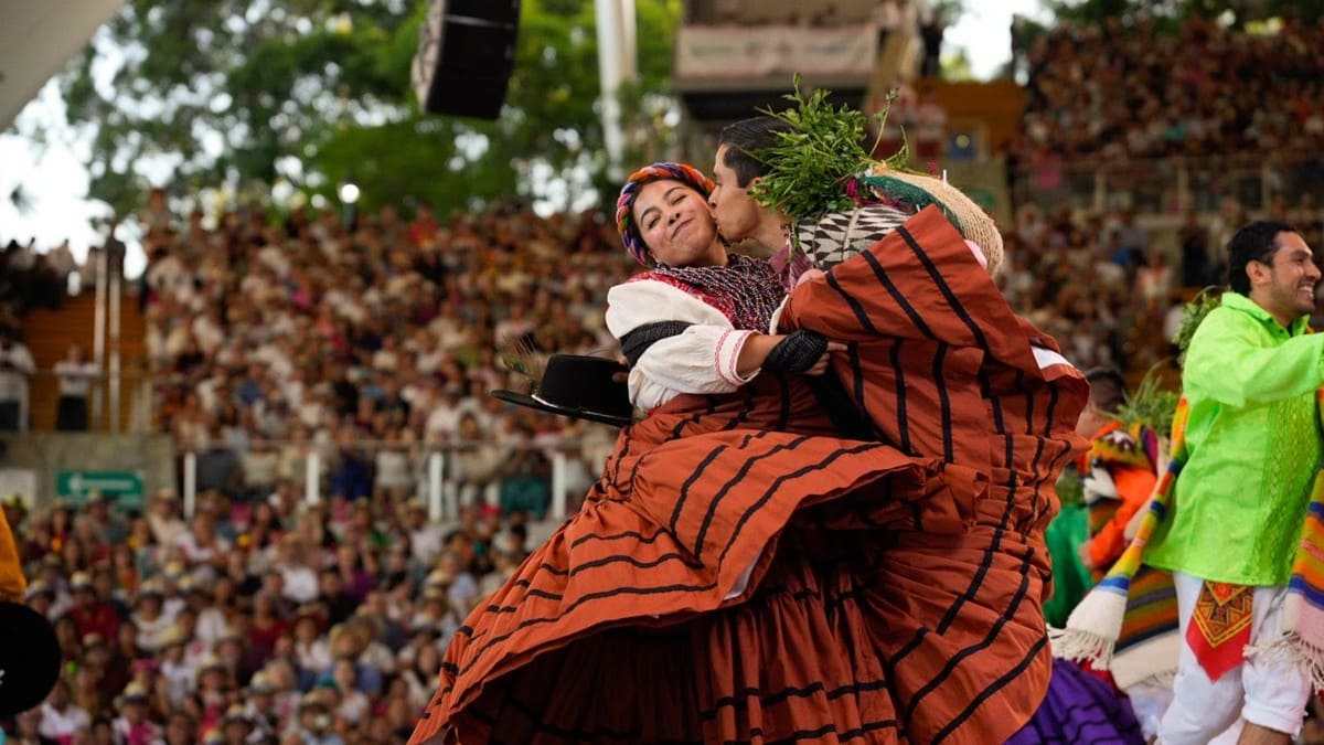 Un hombre y una mujer con ropa regional oaxaqueña bailan y se besan sobre un escenario frente a un gran auditorio