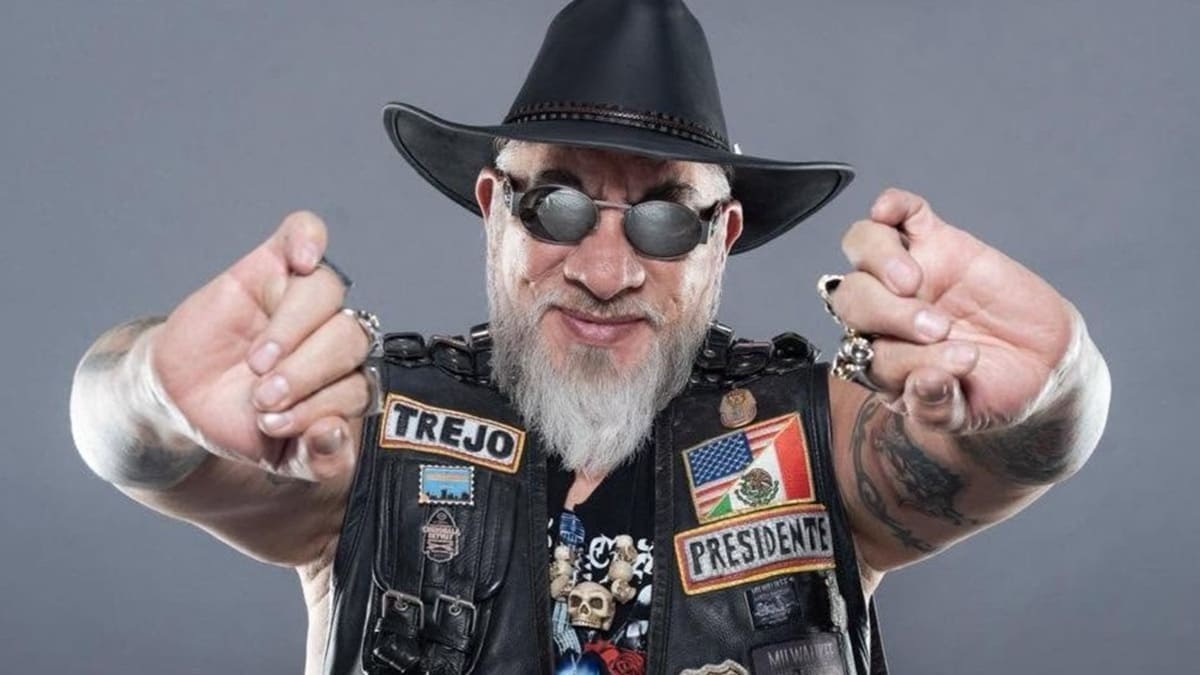 Carlos Trejo con lentes, sobrero, chaleco y saludando a la cámara sonriente