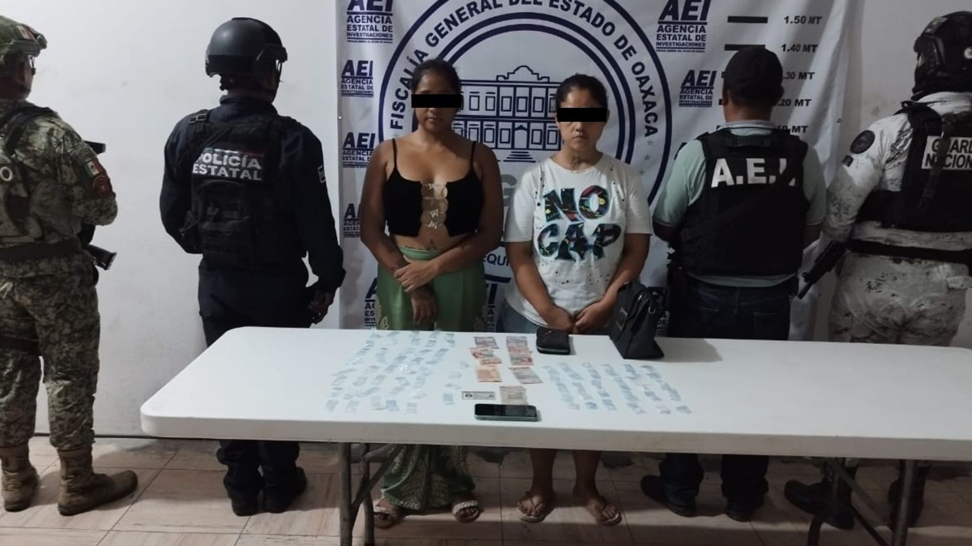 Dos mujeres con el rostro protegido están de pie custodiadas por policías frente a una mesa larga.