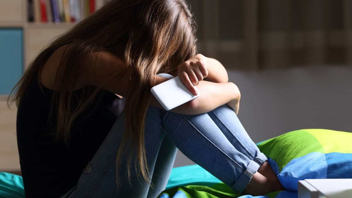 Una joven sentada en la cama tapándose la cara y sosteniendo un celular