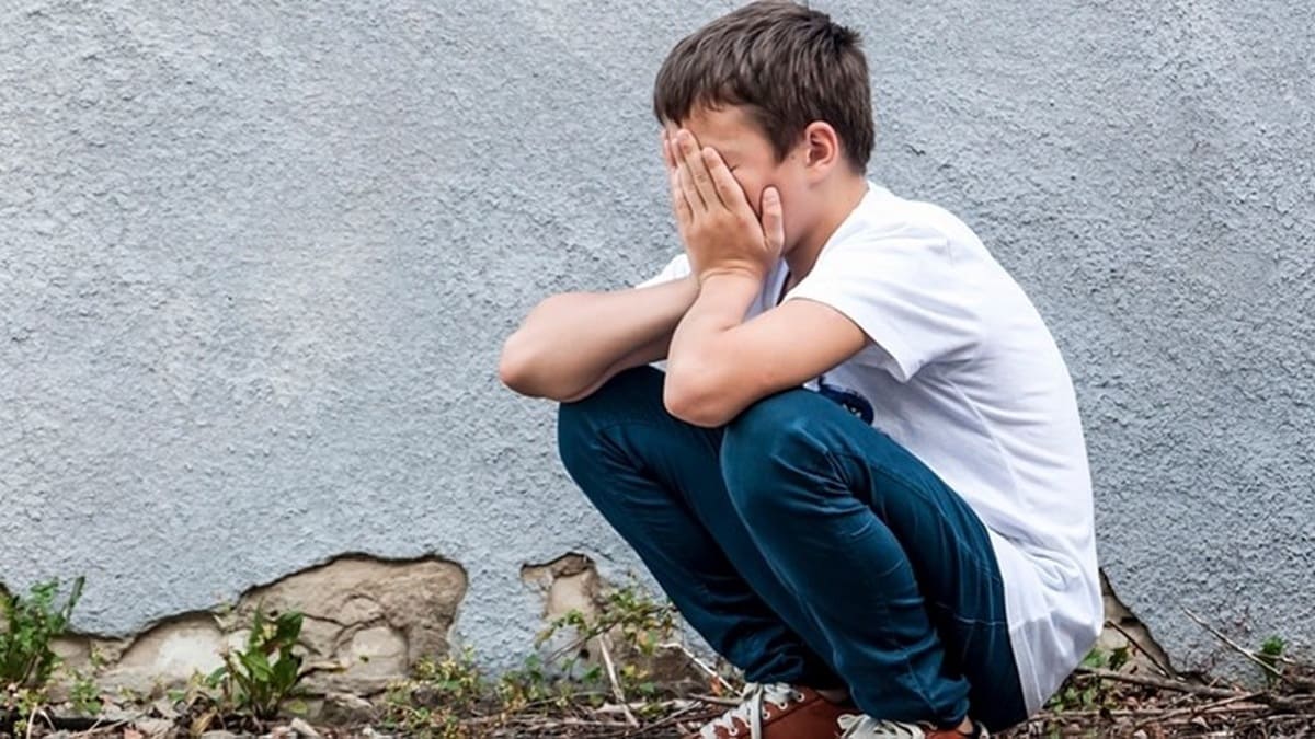 Un niño en cuclillas, usando pantalón de mezclilla y playera blanca, sostiene sus manos contra su rostro en un patio
