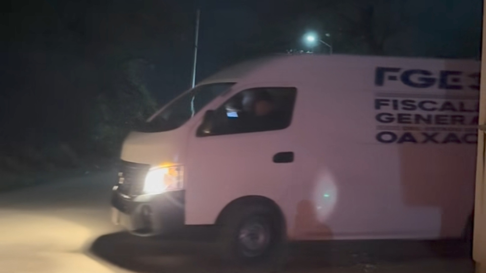Camioneta blanca de la Fiscalía de Oaxaca saliendo de un domicilio con luces encendidas durante la noche.