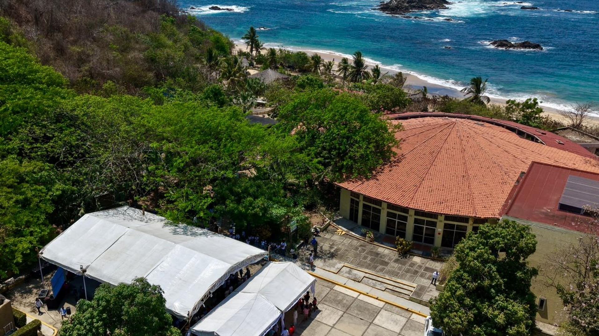 Vista aérea de una playa con arena y un edificio cercano, mostrando la belleza del paisaje costero.