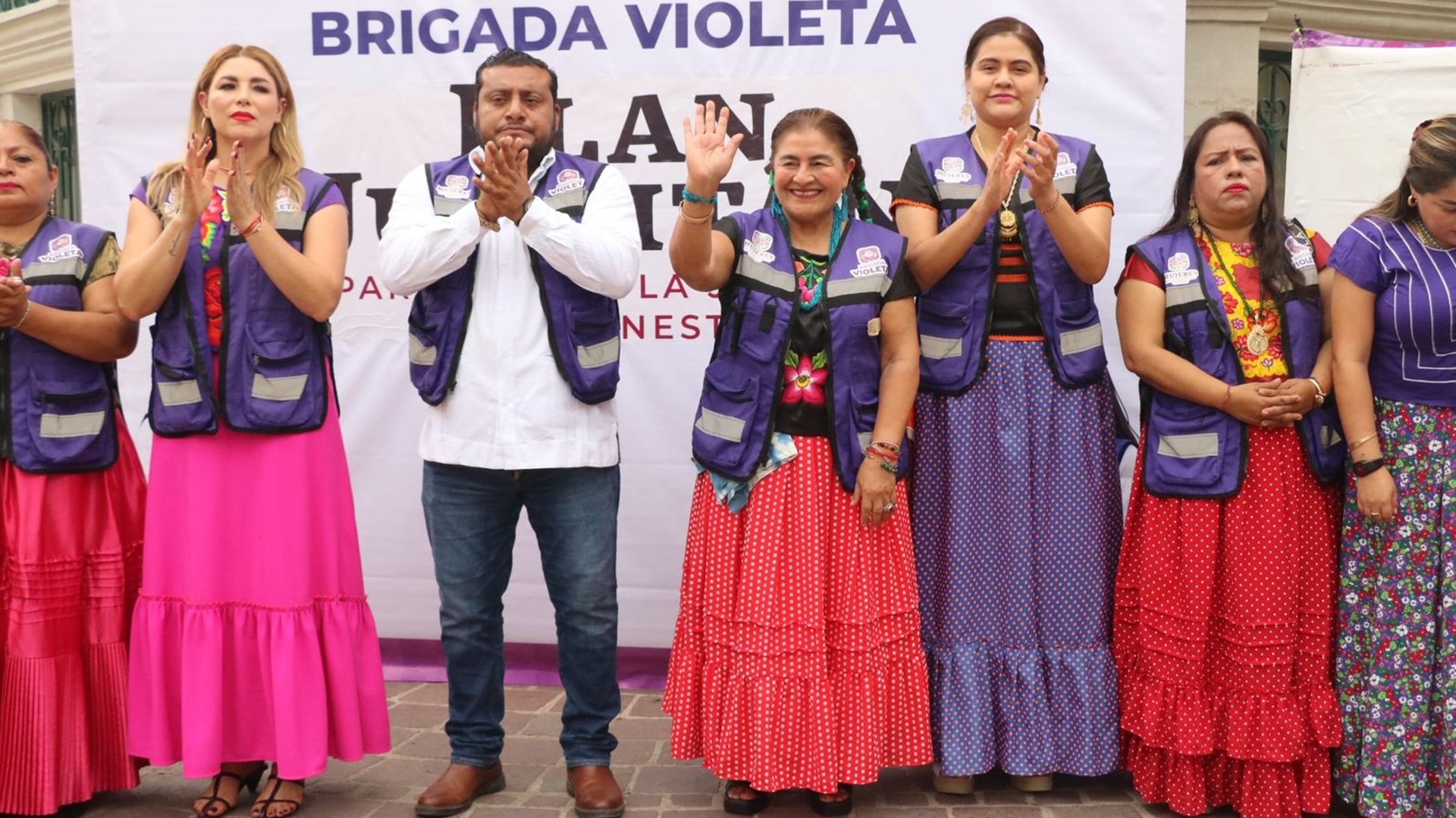 Varias mujeres vestidas con atuendos típicos y un chaleco morado aplauden junto a un hombre también con chaleco morado.