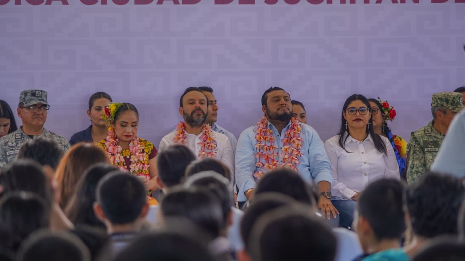 Varias personas con collares de flores sentadas contra una pared y mirando a un auditorio de jóvenes que tienen delante.
